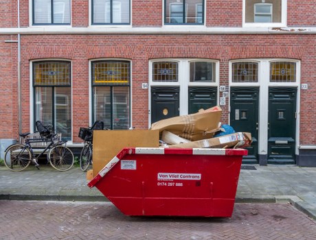 A large red refuse container with a white and red label positioned on a pavement in front of a row of three dark green front doors with white window frames and decorative glass panels in a residential area. The container, used for rubbish collection, is filled with various discarded items including cardboard boxes, paper, and plastic packaging, some of which are spilling over the top. To the left of the container, two bicycles are parked and secured to a metal railing, and a wood panel leans against the railing. The background features a brick terraced building with windows and a small white sign mounted between the two middle doors. The setting appears to be an urban street, likely in Fitzrovia, with a mix of residential and rubbish storage spaces, aligning with the services of Garden Clearance Fitzrovia for rubbish removal.