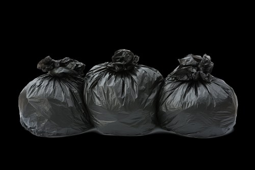 Three large black plastic rubbish bags, tightly knotted at the top, are lined up on a dark surface with a solid black background. The bags appear full and are made of glossy, slightly crinkled plastic material, showing subtle reflections of light on their surfaces. The bags are positioned in a row, with the middle bag slightly behind the others, indicating they are resting on a flat, smooth surface. The environment suggests an outdoor setting, possibly on a driveway or pavement, linked to a garden clearance service in Fitzrovia. This image effectively illustrates generic rubbish collection and disposal tasks, which Garden Clearance Fitzrovia manages as part of their rubbish removal services in the local area.