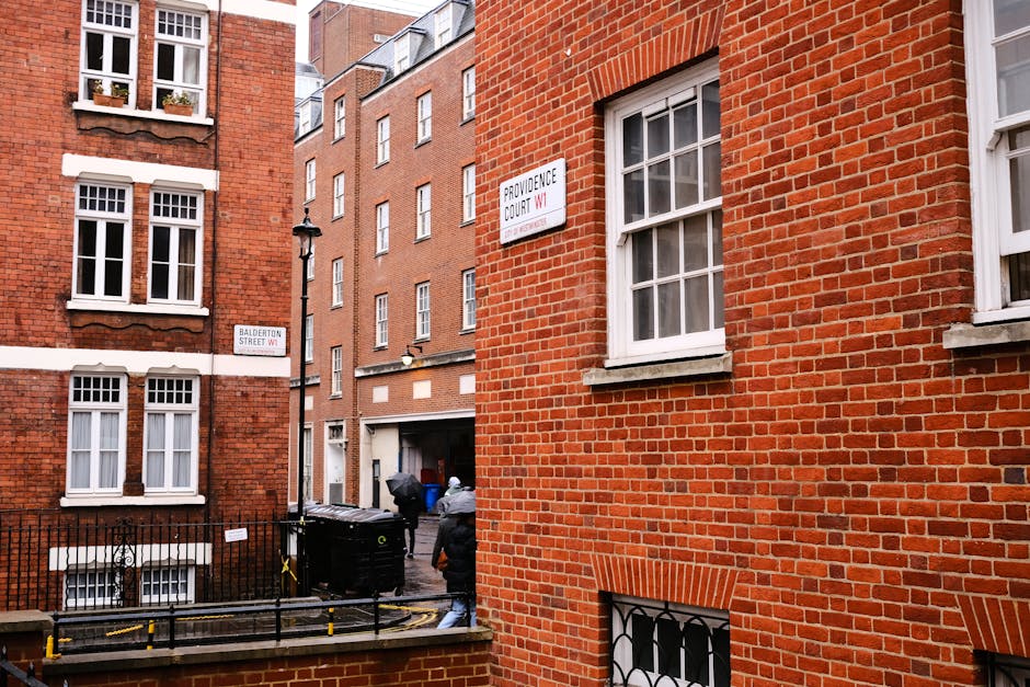 The image depicts a narrow urban alleyway or pedestrian path situated between closely positioned residential buildings in a city. On the left side, there is a brick wall with a built-in wooden planter