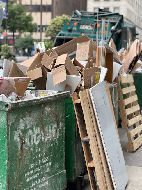 A large green wheeled rubbish bin filled with bags of garden or construction waste, placed outdoors against a dark brick wall. The bin contains several red and white bags labeled with gardening or bui
